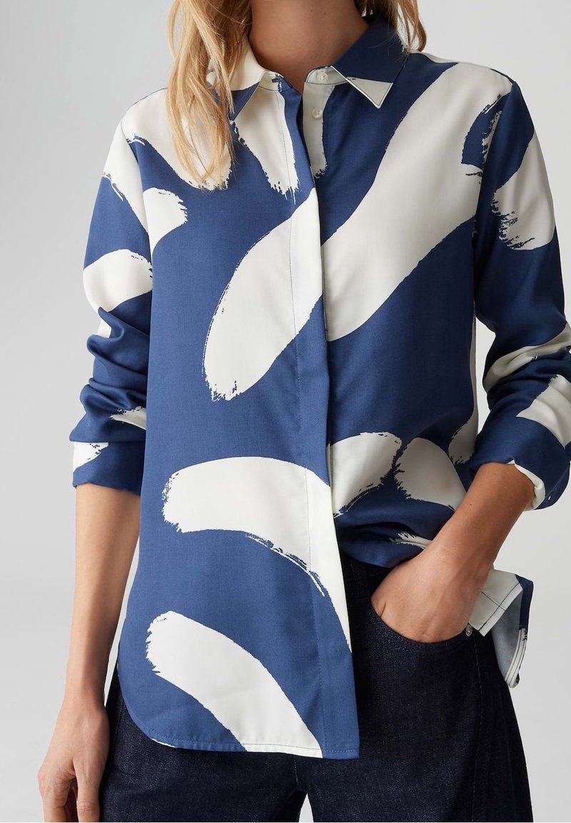 Femme portant une chemise bleue à boutons avec de grands motifs abstraits de coups de pinceau blancs et un pantalon foncé, une main dans la poche.