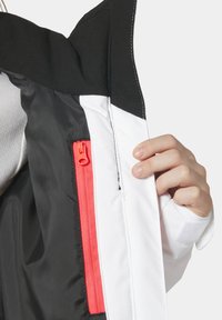 Izolovaná bunda s černým límcem a bílým tělem. Obsahuje oranžový zip a zapínání na suchý zip na rukávech. Hladká a texturovaná tkanina.