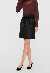 Jupe en cuir noir avec une taille cintrée et un détail de ceinture, longueur genou, associée à un haut à manches longues bordeaux et des talons noirs.