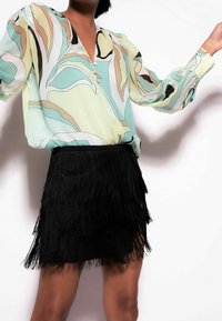 Lys grøn bluse med et flerfarvet bladmønster, der har en V-udskæring og tre knapper. Parret med en sort fringed mini-nederdel.