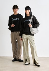 Twee modellen dragen bijpassende zwarte sweatshirts met grafische letters; één paar heeft lichtbeige cargobroeken met ritsen en zakken.