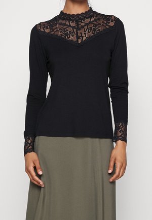 Czarna bluzka z długim rękawem z koronkowymi detalami na biuście i mankietach, noszona z oliwkowozieloną spódnicą, pokazana od szyi do bioder na białym tle.