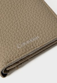 Calvin Klein piniginė iš tekstūruoto taupe spalvos odos, su siuvinėtomis kraštinėmis ir sidabriniu iškilminiu logotipu. Sulanksta dizainas su lygiu vidumi.