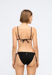 Black bikini with thin straps and scalloped edges. Features a tie back closure and a small logo on the rear. Smooth texture.