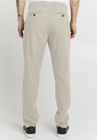 Beige Hosen mit geradem Bein, ausgestattet mit zwei hinteren Taschen mit schwarzen Knöpfen und einem glatten, strukturierten Stoff. Darunter schwarze Sneaker.