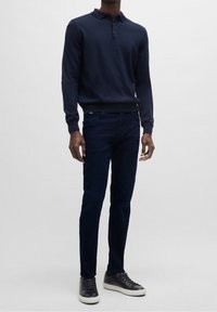 Man i mörk marinblå långärmad polo, mörkblå jeans och svarta sneakers med vita sulor, står mot en enkel ljus bakgrund.