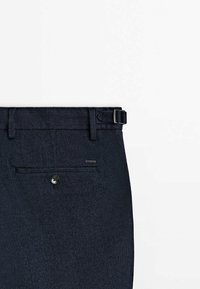 Mörkblå herringbone-byxor med bälteshällor, knappstängning och en bakficka med en knapp samt en liten m ärkesetikett.