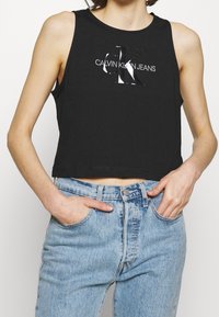 Schwarzes, kurzes Tanktop mit einem glänzenden silbernen "CK"-Logo und dem Text "CALVIN KLEIN JEANS", kombiniert mit hellblauen Jeans.