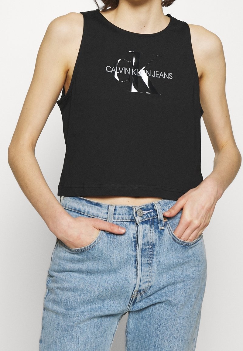 Schwarzes, kurzes Tanktop mit einem glänzenden silbernen "CK"-Logo und dem Text "CALVIN KLEIN JEANS", kombiniert mit hellblauen Jeans.
