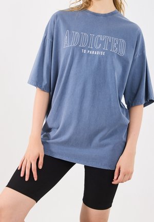 Person iført en oversized blå T-shirt med teksten "ADDICTED TO PARADISE" parret med sorte cykelshorts mod en ensfarvet baggrund.