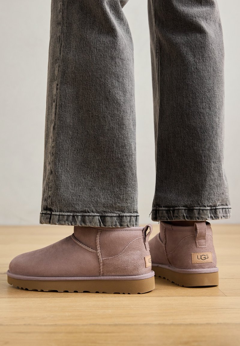 Stivali ankle in suede rosa con punta arrotondata e tab di estrazione posteriore, abbinati a jeans in denim grigio cropped, su un pavimento in legno.