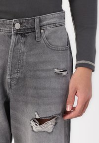 Jean en denim gris avec des déchirures effilochées au genou, design classique à cinq poches, et détail en bouton argenté à la taille.
