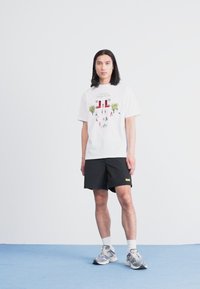 Hvit t-skjorte med fargerik grafisk trykk som viser trær og figurer, sammen med svarte shorts. Modellen står på blått teppe mot en ensfarget bakgrunn.