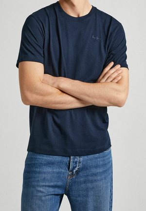 Dunkelblaues Baumwoll-T-Shirt mit kurzen Ärmeln, rundem Ausschnitt und dezentem Logo-Stick auf der Brust. Kombiniert mit hellblauen Jeans.