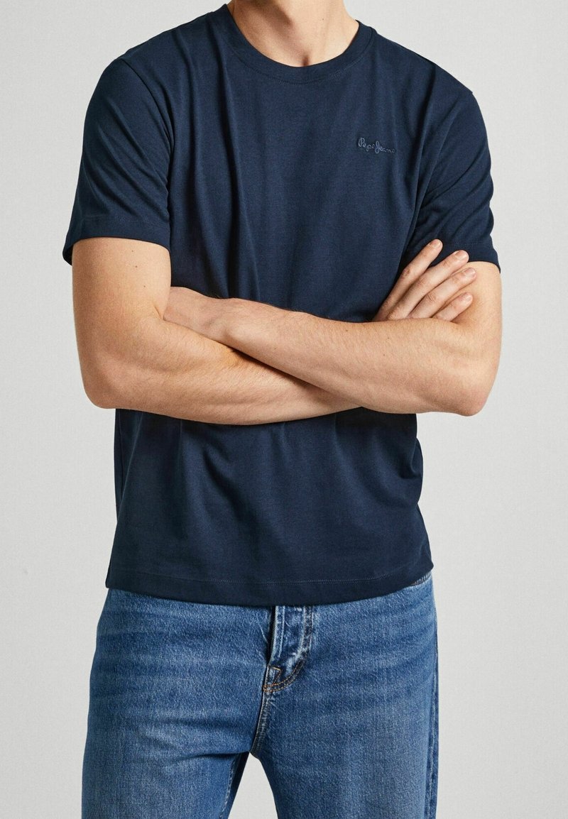 Camiseta de algodón azul marino con mangas cortas, escote redondo y un sutil bordado del logo en el pecho. Combinada con jeans de color azul claro.