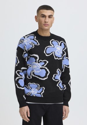 Ung mand iført en sort sweater med store lyseblå og hvide abstrakte blomstermønstre, stående mod en ensfarvet baggrund.