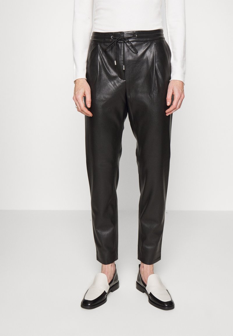Pantalons en cuir noir ajustés avec plis à l'avant, taille à cordon, et texture lisse. Associés à des mocassins noirs et blancs.
