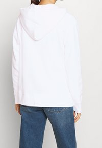 Persona con una sudadera blanca lisa y vaqueros azules de espaldas contra un fondo neutro.