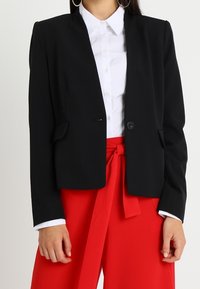 Kvinna iförd en svart kavaj över en vit skjorta med knappar, i kombination med högmidjade röda byxor som är knutna med ett tygbälte.