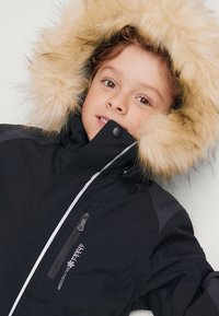 Zwarte winterjas met een met bont gevoerde capuchon, voorzien van een rits aan de voorkant en zijzakken, geaccentueerd met witte piping en logo-detail.