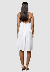 Vestido de verano blanco en capas con tirantes finos ajustables y tejido texturizado. El dobladillo cae por encima de la rodilla, combinado con zapatos de tacón desnudos.