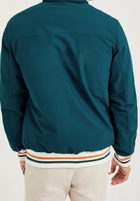 Teal bomberjacka med en slät yta, med ribbade ärmar och nederkant i vita, gröna och orangea ränder. Baksidan visar axelsömmarna.