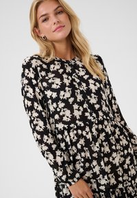 Mustapituinen pitkähihaisen mekko, jossa on valkoinen kukkakuosi, pleat-detaljinen muotoilu, nappikiinnitys ja pehmeä, teksturoitu kangas.