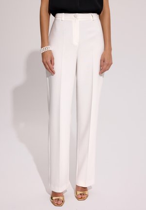 Kobieta ubrana w białe, wysokowęściowe, dopasowane spodnie z przednią plisą, czarny top, perłowa bransoletka i złote sandały na obcasie, stojąca na tle o jednolitym kolorze.