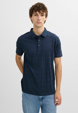 Joven con una camisa polo azul marino de manga corta y vaqueros azul claro, de pie frente a un fondo liso gris claro.