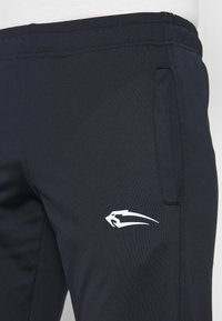 Nahaufnahme von marineblauen Sporthosen mit Seitentasche und stilisiertem weißen Logo am oberen Oberschenkel.