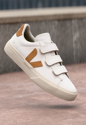 Baskets blanches avec trois bandes Velcro et détail de logo en daim marron, flottant au-dessus d'une surface pavée avec un arrière-plan flou.