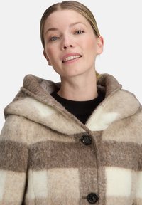 Fleecejacke mit Kapuze, die ein wechselndes braun- und cremefarbenes kariertes Muster aufweist, zwei schwarze Knöpfe hat und aus weichem, strukturiertem Stoff besteht.