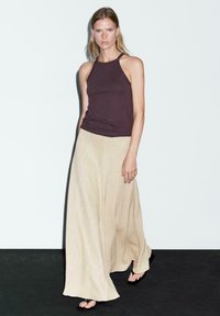 Top senza maniche marrone abbinato a una lunga gonna beige fluente con una sottile trama a spina di pesce. Modella in piedi su una superficie scura.