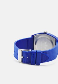 Montre en caoutchouc bleu avec un bracelet texturé, boucle en acier inoxydable et un dos de boîtier transparent montrant un logo. Cadran carré avec un design simple.