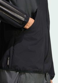 Crna jakna od glatkog materijala s džepom na zip. Ima reflektirajuće trakice na rukavima i blago teksturirani donji dio.