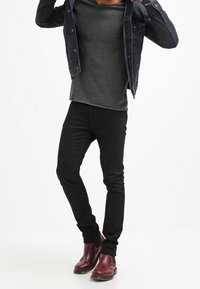 Veste en denim noir sur une chemise à manches longues grise, associée à un pantalon noir ajusté et des bottines rouges foncées. Design simple, coupe slim.