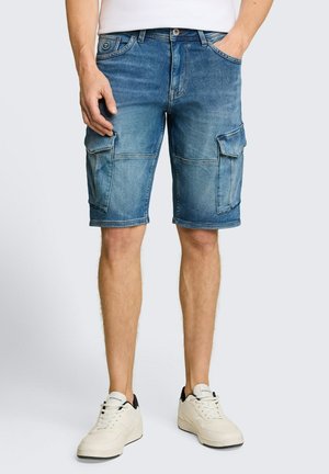 Mężczyzna ubrany w niebieskie dżinsowe szorty typu cargo o długości do połowy uda, białe trampki i białą koszulę, z jedną ręką w kieszeni.