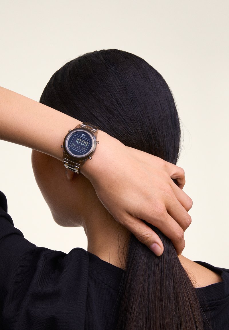 Orologio digitale con quadrante rotondo nero, hardware argentato e bracciale metallico. Mostra l'ora e la data. Posizionato su un polso con capelli scuri nei dintorni.