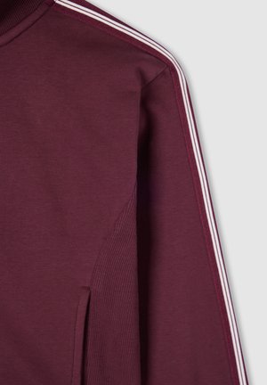 Indumento a maniche lunghe di un rosso scuro con tre strisce bianche che scorrono lungo la spalla e la cucitura della manica, su uno sfondo grigio.