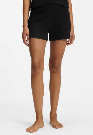 Personne portant un short en maille côtelée noire, debout pieds nus devant un fond blanc uni.