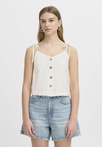 Top blanco corto con tirantes ajustables, que presenta un escote en V y cinco botones redondos. Combinado con pantalones cortos de mezclilla azul claro.