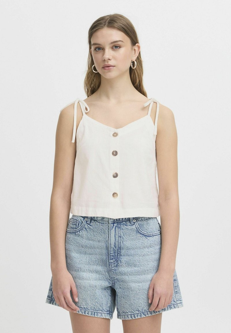 Top blanco corto con tirantes ajustables, que presenta un escote en V y cinco botones redondos. Combinado con pantalones cortos de mezclilla azul claro.