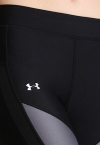 Leggings atletici neri con una texture liscia, caratterizzati da un pannello grigio e dal logo Under Armour sul lato. Primo piano dell'area superiore della coscia.