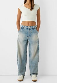 Vrouw in een witte cropped t-shirt, losse lichtblauwe vervaagde jeans met handen in de zakken, en witte sneakers tegen een effen achtergrond.