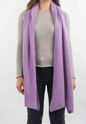 Mujer vistiendo una bufanda larga lavanda sobre un suéter beige de manga larga y pantalones negros, de pie contra un fondo blanco liso.