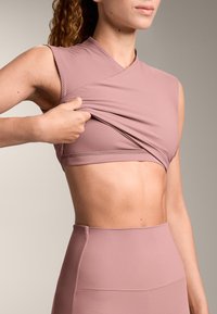 Top cu mâneci scurte, roz, mulat, cu textură nervurată, tras în sus pentru a dezvălui un sutien sportiv asortat, purtat cu colanți roz cu talie înaltă.