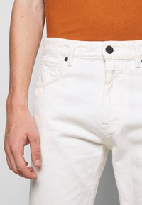 Jeans de mezclilla blancos con una textura suave, detallados con un botón negro y costuras sutiles. Una mano posicionada cerca de la cinturilla es visible.