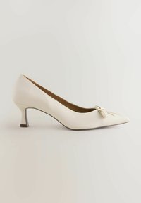 Zapato de tacón blanco de cuero con punta afilada, pequeño lazo decorativo y tacón cuadrado de altura media. Textura suave con un brillo sutil.