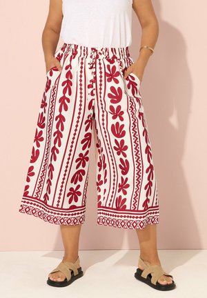 Kvinde iført hvide vide ben-korte bukser med røde blomster- og geometriske mønstre, kombineret med beige sandaler og en hvid top.