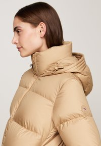 Jachetă puffer bej cu guler înalt și glugă ajustabilă. Prezintă secțiuni matlasate și o etichetă cu logo pe mânecă. Textură netedă.